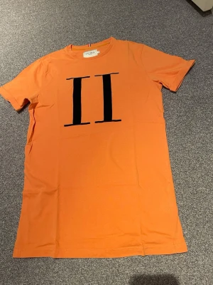 Orange t-shirt från Les Deux - Snygg orange t-shirt från Les Deux med svart tryck av två romerska ettor på bröstet. Klassisk rund hals och korta ärmar. Perfekt för dig som vill sticka ut med en färgstark och stilren look.