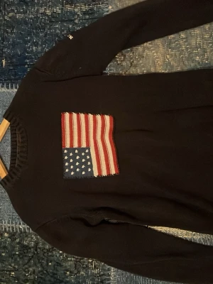 Mörkblå GANT tröja med USA-flagga - Snygg mörsblå  tröja från GANT med en stor broderad amerikansk flagga på bröstet. Tillverkad i 100% bomull och har rund halsringning samt långa ärmar. Perfekt för dig som gillar klassisk och stilren design med en twist.