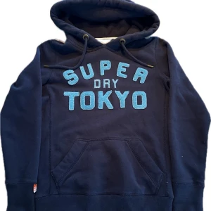 Mörkblå hoodie Superdry Tokyo(extremt sällsynt) - Mörkblå hoodie från Superdry med stor ljusblå text 'SUPER DRY TOKYO' på bröstet. Klassisk känguruficka framtill, justerbar huva med snören och ribbade muddar. Perfekt för en avslappnad streetwear-look. Archive! Hoodien sitter mer slim och är small i passform. Så skulle gå upp en storlek