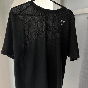 Svart Gymshark t-shirt seamless M - Svart t-shirt från Gymshark med diskret mönstrad mesh upptill och klassisk logga på bröstet. Modellen är kortärmad och har en clean, sportig look. Tillverkad i ett lätt och stretchigt syntetmaterial som andas, perfekt för träning.  Det är två stycken t-shirts en oöppnad och därför osäker hur den ser ut men förmodligen liknande den första. Båda två är helt oanvända. Säljes pga att dem är förstora. Pris går att diskutera vid köp av båda två, kan tänka mig båda för 800 Står ej för frakten. 