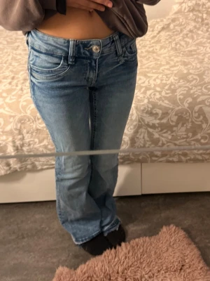 Blå bootcut jeans med låg midja - Säljer ett par ljusblå bootcut jeans med låg midja och klassiska fem fickor från Gina i storlek Young 152. Jeansen har en avslappnad passform och är tillverkade i denim med lätt tvättad look. Perfekta för en casual och trendig stil.