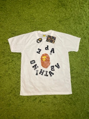 Vit A Bathing Ape t-shirt med tryck - Säljer en vit t-shirt från A Bathing Ape med det klassiska apansikte-trycket i brunt och gult samt texten 'A BATHING APE' i svart runt motivet. T-shirten har rund hals och korta ärmar. Perfekt för dig som gillar streetwear och vill sticka ut.