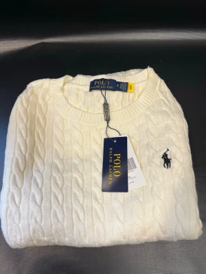 Vit kabelstickad tröja Polo Ralph Lauren - Kabelstickad vit tröja från Polo Ralph Lauren med rund halsringning och den klassiska broderade loggan i marinblått på bröstet. Tröjan har ribbade muddar vid ärmslut och nederkant. Perfekt för en stilren och tidlös look.