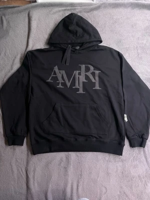 Svart hoodie från Amiri med logga - Snygg svart hoodie från Amiri med stor grå logga framtill. Klassisk känguruficka och huva med dragsko. Tillverkad i mjukt bomullsmaterial och har ribbade muddar vid ärmar och nederkant. Perfekt för en avslappnad och trendig streetwear-look.