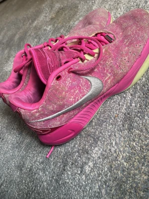 Lebron 21 - Säljer ett par unika rosa Nike sneakers med silverfärgad swoosh och coolt mönstrad ovandel. Skorna har rosa snören, beige detaljer vid snörningen och en ljusgul sula. Perfekta för dig som vill sticka ut med färg och stil.