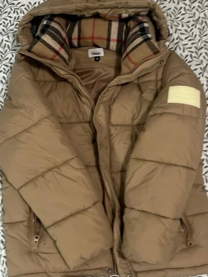Beige pufferjacka från Burberry - Snygg beige pufferjacka från Burberry med klassiskt rutigt foder i huvan och stor patch med Burberry London England på ärmen. Jackan har dragkedja, fickor och en rejäl huva med fluffig känsla. Perfekt för kalla vinterdagar och riktigt trendig look.