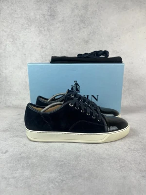 Svarta sneakers från Lanvin - Snygga svarta sneakers från Lanvin med ovandel i sammet och lackad tå. Skorna har vita sulor och svarta skosnören. Perfekta för dig som vill ha en stilren och lyxig look med en twist av materialmix.