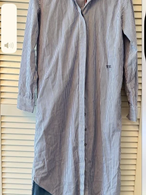 Randig skjorta från Tommy Hilfiger - Klassisk långärmad skjorta från Tommy Hilfiger i vitt och blått med smala vertikala ränder. Skjortan har knappar framtill, krage och diskret logga broderad på bröstet. Tillverkad i bomull för en skön och luftig känsla.