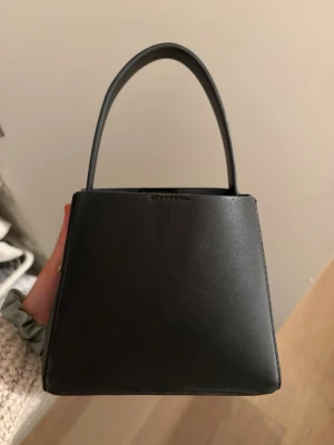 Svart handväska från Gina - Stilren svart handväska från Gina i slätt läder. Väskan har en elegant, fyrkantig form med ett topphandtag och magnetlås. Invändigt finns ett fack med dragkedja för småsaker. Perfekt accessoar för en trendig look. Inget långt band finns.