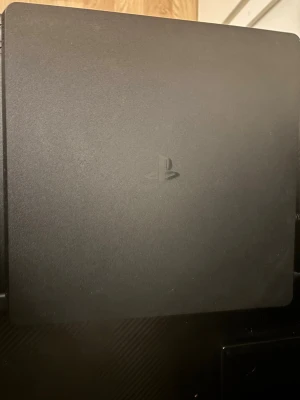 PlayStation 4 Slim spelkonsol svart - Säljer en svart PlayStation 4 Slim från Sony. Konsolen har en stilren, rektangulär design med matt finish och PlayStation-loggan på ovansidan. Materialet är plast och den har anslutningar för HDMI och nätverk på baksidan. Perfekt för gaming och streaming.