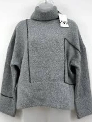 Grå stickad polotröja med svarta detaljer - Trendig grå stickad polotröja med svarta kontrastsömmar och en unik asymmetrisk ficka framtill. Tröjan har en lös passform, långa ärmar och hög krage. Perfekt för dig som gillar en modern och stilren look.