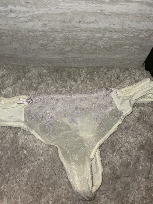 Krämvita spetsiga stringtrosor - Snygga krämvita stringtrosor med transparent mesh och lila broderade blommor framtill. Trosorna har tunna sidor och dekorativ spetskant. Materialet är lätt och stretchigt, perfekt för en feminin look.