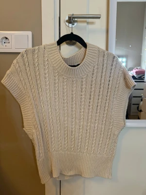 Beige stickad tröja med ribbade detaljer - Snygg beige stickad tröja med bred ribbad kant nedtill och vid halsen. Tröjan har korta ärmar och ett vertikalt kabelstickat mönster som ger en trendig look. 