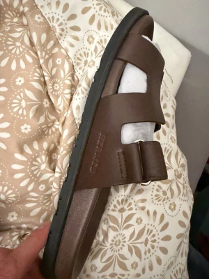 Bruna sandaler från Birkenstock - Snygga bruna sandaler från Birkenstock med två breda remmar i läder och justerbart spänne. Klassisk slip-in modell med svart sula och bekväm fotbädd. Perfekta för en avslappnad och trendig stil.