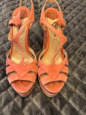 Korallfärgade platåpumps från Miu Miu - Säljer ett par snygga korallfärgade platåpumps från Miu Miu med kilklack och remmar i mocka. Skorna har en cool silvrig detalj längs sulan och öppen tå. Storlek 38 och sulan är i läder. Högsta klackhöjd 15cm. 