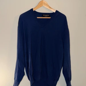 Mörkblå V-ringad tröja YSL - Snygg mörkblå stickad tröja från Yves Saint Laurent med V-ringning. Klassisk och enkel design med långa ärmar och ribbade muddar. Perfekt att matcha med jeans eller chinos för en clean look.