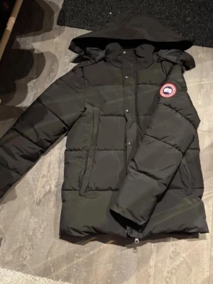 Svart dunjacka från Canada Goose - Säljer en svart dunjacka från Canada Goose med avtagbar huva och pälsdetalj. Jackan har två stora framfickor, dragkedja och tryckknappar samt den klassiska logotypen på ärmen. Perfekt för kalla vinterdagar.