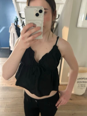 Svart croppad linnetopp med rosett - Snygg svart croppad linnetopp med smala axelband och söt rosettdetalj framtill. Toppen har v-ringning och volangkant vid bysten, vilket ger en feminin look. Perfekt att matcha med jeans eller kjol för en trendig stil.