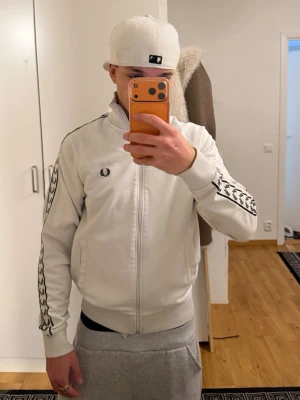 Vit Fred Perry track jacket - Vit track jacket från Fred Perry med svarta laurel-wreath-loggor längs ärmarna och broderad logga på bröstet. Dragkedja framtill, ribbade muddar och krage. Perfekt för en sportig och stilren look.     