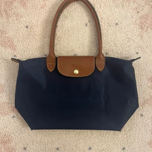 Mörkblå Longchamp Le Pliage väska - Snygg mörkblå handväska från Longchamp Le Pliage med bruna handtag och lock i läder. Väskan är tillverkad i slitstark nylon och har guldfärgad knappdetalj. Klassisk och tidlös design som enkelt kan vikas ihop. I storlek M 