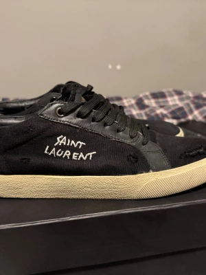 Svarta Saint Laurent sneakers - Svarta sneakers från Saint Laurent med vit broderad logga på sidan. Skorna har svart canvas och läderdetaljer, svarta skosnören och en beige sula. Snygg, avslappnad design med vintagekänsla och diskreta slitningar för en cool look. Hål i sulorna som du kan se på sista bilden kom gärna med förslag.