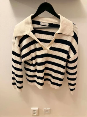 Randig stickad tröja från MNG - Snygg stickad tröja från MNG med breda svarta och vita ränder. Tröjan har en djup V-ringning och stor krage som ger en trendig look. Långa ärmar och mjukt material, perfekt för lager-på-lager-stil.