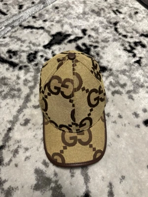 Gucci beige keps med GG-mönster - Säljer en beige keps från Gucci med det klassiska bruna GG-monogrammet över hela kepsen. Kepsen har bruna detaljer och är tillverkad i en mix av merinoull och kashmir. Snygg och tidlös accessoar för dig som gillar lyxiga märken.