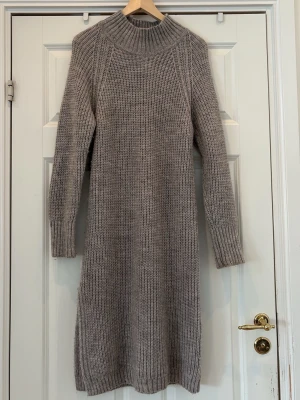 Grå stickad klänning Levate Room - Säljer en grå stickad klänning från Levate Room i storlek M. Klänningen har polokrage, långa ärmar och ribbstickade detaljer vid hals, ärmslut och nederkant. Perfekt för lager på lager och riktigt mysig under kalla dagar. Klänningen har en kort slits på ena sidan.