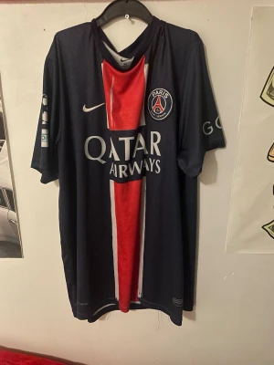 PSG matchtröja Nike mörkblå - Snygg mörkblå Paris Saint-Germain matchtröja från Nike med röd och vit rand i mitten, PSG-logga på bröstet och Qatar Airways som sponsor. Tillverkad i lätt och ventilerande material, perfekt för fotboll eller träning. Korta ärmar och klassisk passform.