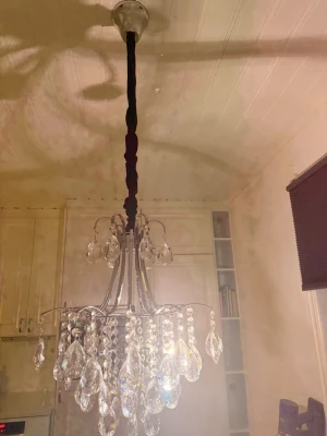 Klassisk kristallkrona i silver - Säljer en elegant kristallkrona med silverfärgad metallstomme och hängande klara prismor i olika former. Lampan har en lyxig känsla och ger ett snyggt ljusspel i rummet. Perfekt för dig som vill ha något extra i taket. Svart sammets tyg längs kablarna ger en mer lyxig och modern känsla. Köpt 2025 så i nyskick. Djur och rökfri hem