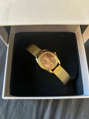 Guldfärgad mesh-klocka med vinröd urtavla - Stilren klocka från guldfynd med guldfärgat mesh-armband och vinröd urtavla. Urtavlan har romerska siffror och boetten är rund med räfflad kant. Perfekt accessoar för dig som vill ha en elegant och trendig look.