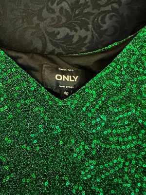 Grön linne  från ONLY - Snygg grön linne  från ONLY täckt med glittrande paljetter. Klänningen har smala axelband och V-ringning, perfekt för dig som vill sticka ut. Materialet är glansigt och skimrande, vilket ger en festlig look.