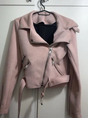 Ljusrosa mockajacka från zara - Snygg ljusrosa bikerjacka med flera silvriga dragkedjor, bälte i midjan och bred krage. Jackan har lång ärm med dragkedja vid ärmslut och är tillverkad i ett mjukt material som liknar mocka. Perfekt för att lyfta din outfit. Använd några få gånger och kan bäras från storlekarna Xss-S💗