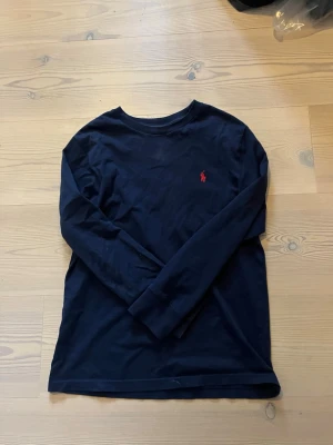 Mörkblå långärmad tröja Polo Ralph Lauren - Klassisk mörkblå långärmad tröja från Polo Ralph Lauren med rund halsringning och den ikoniska röda logotypen broderad på bröstet. Tillverkad i mjuk bomull. Strl 10-12år 