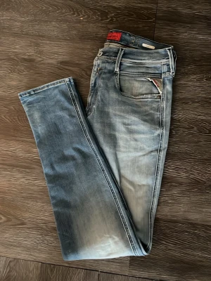 Blå slitna jeans från Replay - Säljer ett par blå jeans från Replay med snyggt slitna detaljer och klassisk femficksmodell. Jeansen har raka ben och ljus tvätt med kontrastsömmar. Märkespatch i läder baktill och Replay-logga på fickan framtill. Perfekta för en avslappnad stil.