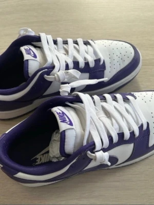 Nike Dunk Low vit/lila sneakers - Säljer ett par Nike Dunk Low sneakers i vitt och lila. Skorna har klassisk låg siluett, vita skosnören och perforerad tåbox för extra ventilation. Perfekta för dig som gillar streetwear och vill sticka ut. Använda 1-2 gånger. 