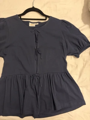 Mörkblå blus med fast knyt - Mörkblå topp med puffärmar och tre dekorativa knyt framtill. Lös och bekväm passform med lätt utsvängd nederdel. Mjukt och skönt material. Passar både till vardag och finare tillfällen.         Aldrig använt.  📦 Snabb frakt 