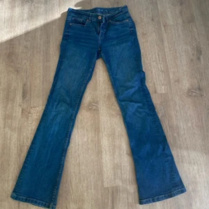 Blå bootcut jeans med normal passform - Snygga blå jeans med bootcut-silhuett och klassisk femficksdesign. Jeansen har normal passform och är tillverkade i ett tåligt denimtyg. Perfekta för dig som gillar en retrovibe och vill ha ett par jeans som sticker ut lite extra.