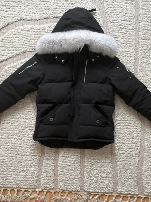 Svart pufferjacka med päls från Moose Knuckles - Säljer en svart pufferjacka från Moose Knuckles med stor huva och lyxig gråvit fuskpäls. Jackan har flera fickor med dragkedjor och tryckknappar samt den ikoniska metall-loggan på ärmen. Perfekt för kalla vinterdagar och riktigt snygg streetstil. Kom dm för fler bilder