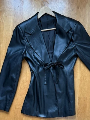 Svart figursydd jacka i skinimitation med knytband - Snygg svart skinnjacka med markerade axlar, breda slag och knytband i midjan för en figurnära siluett. Jackan har lång ärm och slät finish utan synliga fickor eller dragkedjor. Perfekt för dig som vill ha en stilren och trendig look. Liten slitage på axel.