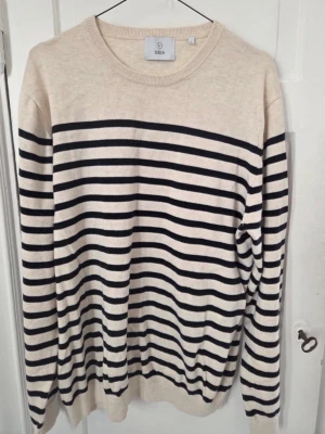 Randig stickad tröja från Bläck - Säljer en beige stickad tröja från Bläck med svarta horisontella ränder. Tröjan har rund halsringning och ribbade muddar vid ärmslut och nederkant. Perfekt för dig som gillar klassisk och stilren design.