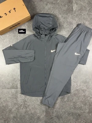 nike miller - Snyggt svart träningsset från Nike med jacka och byxor. Jackan har huva, dragkedja och två fickor, samt reflekterande Nike-logga. Byxorna har resår i midjan och matchande logga. Perfekt för löpning eller träning. Materialet är lätt och andas. Passar 160-170cm