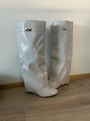 Ljusa vida stövlar i skinnimitation - Säljer ett par ljusgrå, vida stövlar i skinnimitation med slouchy passform och rund tå. Stövlarna har en dekorativ metall-detalj upptill och saknar klack. Perfekta för dig som gillar en trendig och unik look.