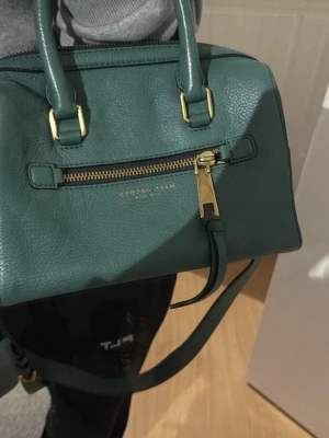 Grön handväska från Marc Jacobs - Snygg grön handväska från Marc Jacobs i strukturerat läder. Väskan har guldfärgade metalldetaljer, dubbla handtag och en avtagbar axelrem. Framtill finns en dragkedjeficka och diskret logga. Perfekt storlek för det viktigaste och ger en cool touch till din outfit.