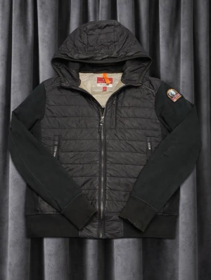 Parajumper cardigan  - Snygg svart pufferjacka från Parajumpers med huva och dragkedja framtill. Jackan har quiltad kropp, mjuka ärmar och ribbade muddar. Märkets patch på ena ärmen och två dragkedjefickor. Perfekt för kalla dagar.