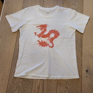 Vit t-shirt med dean blunt "zushi" - Vit t-shirt i 100% bomull med ett stort röd/orange motiv av dean blunt på framsidan. Klassisk rund hals och korta ärmar. Perfekt för dig som gillar coola prints och vill sticka ut med en unik design.