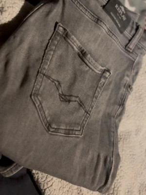 Grå jeans från Replay - Snygga grå jeans från Replay med klassisk femficksdesign och diskreta sömmar på bakfickan. Jeansen har rak passform och är tillverkade i mjukt denimtyg som ger en avslappnad look. Perfekta för dig som gillar stilrena och tidlösa plagg.  Storlek 32 34