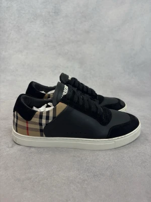 Burberry svarta sneakers med rutigt mönster - Snygga svarta sneakers från Burberry med klassiskt beige, svart och rött rutmönster på hälen. Skorna har ovandel i läder och mocka, vita sulor och svarta skosnören. Perfekta för dig som vill ha en stilren och exklusiv look.