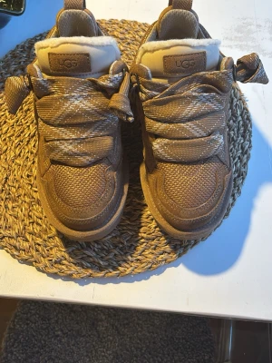 Bruna sneakers från UGG med fuskpäls - Snygga bruna sneakers från UGG med grova mönstrade sulor och breda snören. Ovandelen är i textil och mockaimitation, och insidan har mjuk fuskpäls för extra komfort. Perfekta för dig som vill ha både stil och värme i ett par sneakers.