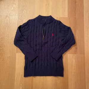 Mörkblå kabelstickad tröja Polo Ralph Lauren - Mörkblå kabelstickad tröja från Polo Ralph Lauren med halv dragkedja och röd broderad logga på bröstet. Tröjan har ribbade muddar och hög krage, perfekt för lager på lager. Klassisk och tidlös design i mjukt material.